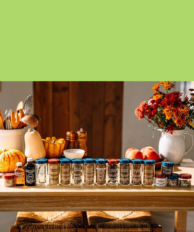 Morton & Bassett premium spices and baking extracts arranged on a wooden kitchen table surrounded by fall pumpkins, peaches, and colorful autumn flowers.