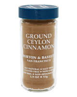 A jar of Morton & Basset Ground Ceylon Cinnamon spice against a solid black background. The clear glass jar shows the warm brown cinnamon powder inside, with white text on the label and a dark blue screw-top lid.