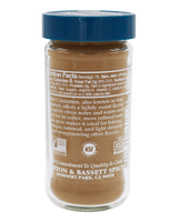 A clear glass spice jar filled with warm brown cinnamon spice powder, featuring a blue screw-top lid and a white label with text and certification logos.