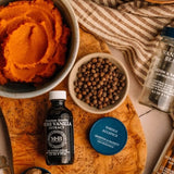 Vanilla extract bottle, bowl of orange ice cream, and container of whole allspice on a wooden surface.
