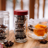 Jar of star anise with a red lid on a wooden surface, surrounded by star anise pods and a glass container with orange slices.