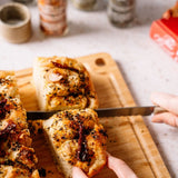 Person cutting a piece of bread with a knife on a wooden cutting board.