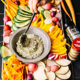 Platter of sliced vegetables with a bowl of dip and seasoning bottles.