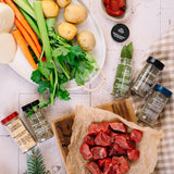 Raw beef chunks on a wooden board with vegetables, spices, and herbs on a checkered tablecloth.