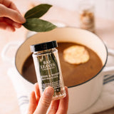 Hand holding a container of Bay Leaves with a bowl of soup in the background