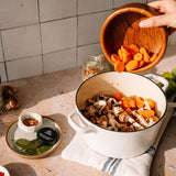 Person adding carrots to a pot of mushrooms on a kitchen counter.