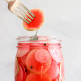 Glass jar with pickled radishes, a fork, and a watermelon slice on a light gray background