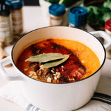 White pot filled with a hearty stew on a wooden table with spices in the background