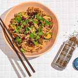 Bowl of noodles with vegetables and a bottle of Caraway Seeds on a white surface