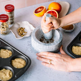 Person using a mortar and pestle on a kitchen counter with ingredients and baking trays.