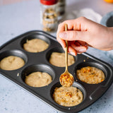 Hand using a spoon to add seasoning to muffin batter in a baking tray.