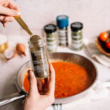 Person adding celery seed to a jar with a pan of red sauce in the background