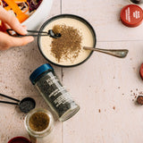 Person adding spices to a bowl of creamy dish on a textured surface with a jar of spices.