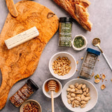 Spices, nuts, and a honey dipper on a gray surface with a wooden cutting board.