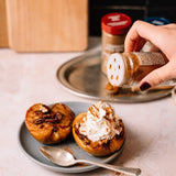 Person sprinkling cinnamon on a dessert with whipped cream, surrounded by coffee jars.