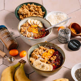 Two bowls of a breakfast dish with bananas, oranges, and various toppings on a tiled surface.