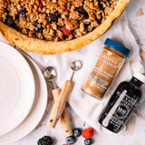 Baked pie with cinnamon and vanilla extract, berries, and measuring spoons on a white surface.