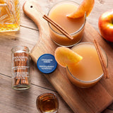 Two glasses of apple cider with cinnamon sticks and apple slices on a wooden cutting board, surrounded by cinnamon sticks and apples.