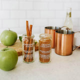 Two jars of Morton & Bassett cinnamon sticks and ground cinnamon on a table with green apples and copper mugs.