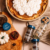 Pumpkin pie with whipped cream on a table with spice bottles and ingredients.