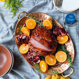 Decorative platter with sliced ham, oranges, and pomegranates on a blue checkered tablecloth.
