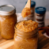 Jar of brown sugar scrub with a wooden spoon lifting some scrub above it, on a wooden surface.