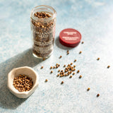 Jar of Coriander Seed with a small dish and scattered seeds on a light blue surface