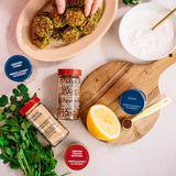 Spices, herbs, and a lemon on a table with a bowl of food and a plate.