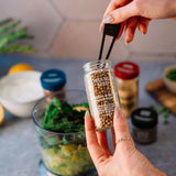 Person holding a jar of 'Coriander Seed' seasoning with a kitchen counter in the background