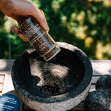 Hand pouring pepper into a stone mortar with a blurred green outdoor background