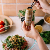 Person adding cumin from a container into a dish with vegetables on a kitchen counter.