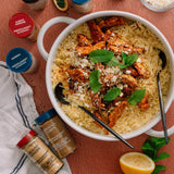 Bowl of pasta with chicken and herbs, surrounded by spice containers on a red surface.