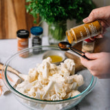 Person seasoning cauliflower with curry powder in a kitchen setting