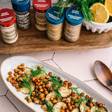 Spice jars on a wooden shelf with a dish of roasted chickpeas and zucchini in the foreground.
