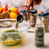 Person adding dill weed to a bowl with other ingredients on a kitchen counter.