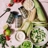 Assorted ingredients including corn on the cob, radishes, lime, and various seasonings on a pink surface.