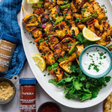 Platter of grilled chicken skewers with lemon wedges, herbs, and a small bowl of yogurt dip, accompanied by spices on a white surface.