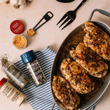 Seasoned chicken breasts in a pan with spices and utensils on a table.