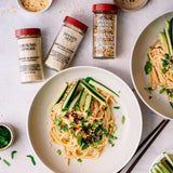 Spaghetti dish with cucumber slices and herbs, surrounded by seasoning jars on a light background.
