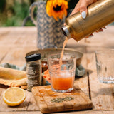 Person pouring a drink from a gold thermos into a glass on a wooden table with a sunflower in the background.