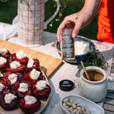 Person holding a bottle of herbs from Provence seasoning over a dish with plums and cheese.