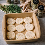 A square baking dish with nine uncooked herb biscuits on parchment paper, surrounded by various jars of dried herbs and fresh herbs on the counter.