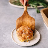 A hand holds a wooden spatula placing a round biscuit with herbs and salt onto a small white plate.