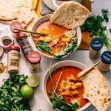 Two bowls of vibrant curry soup with naan, surrounded by fresh cilantro, lime, and spice jars, create a flavorful meal setting.