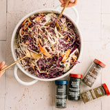 Hands mixing colorful coleslaw in a bowl with poppy, sesame, mustard, and celery seeds on the side for seasoning.