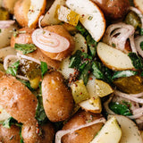 Close-up of a potato salad with sliced red potatoes, pickles, shallots, and herbs, seasoned with mustard seeds and spices.
