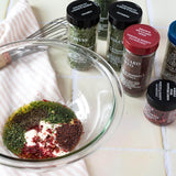 Glass bowl with herbs, mustard seeds, and spices on a striped cloth. Spice jars labeled chives, parsley, and peppercorns nearby.