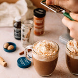 Two mugs of coffee topped with whipped cream are garnished with grated nutmeg. Spices and ingredients are scattered around.