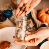 Hands hold a jar of whole nutmeg with a pumpkin and spices in the background, creating a warm, autumnal kitchen scene.
