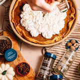 Pumpkin pie topped with whipped cream, surrounded by spices like cloves, nutmeg, and allspice, on a striped cloth.